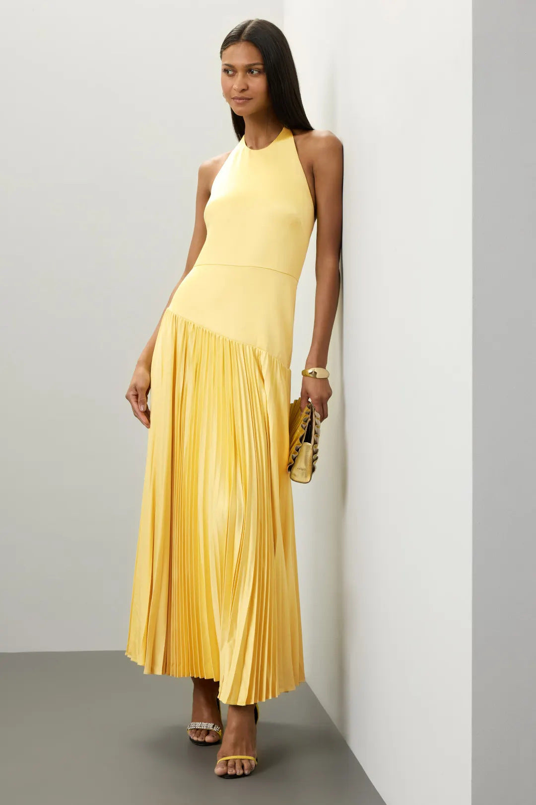 Woman wearing a yellow pleated halter dress for summer with sandals and a matching clutch bag